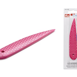 Könnte beinhalten: Ein rosa Eck- und Kantenformer mit weißen Punkten. Das Werkzeug hat ein spitzes Ende und einen abgerundeten Griff mit einem kleinen Loch. Die Verpackung trägt die Aufschrift "Frym Love" und "Corner and edge shaper".
