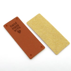 Puede incluir: Dos etiquetas rectangulares de cuero. Una es marrón con las palabras "hand made with" y un corazón en relieve. La otra etiqueta es de color beige claro. Ambas etiquetas tienen cuatro pequeños agujeros en cada esquina.