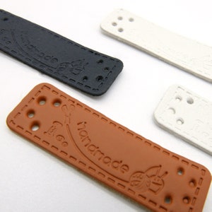 Op de afbeelding: Close-up van vier rechthoekige leren labels in zwart, bruin en wit. Elk label heeft het woord "handmade" in reliëf, samen met decoratieve stiksels en voorgeboorde gaten om te naaien of te bevestigen. De labels zijn voor knutselen.