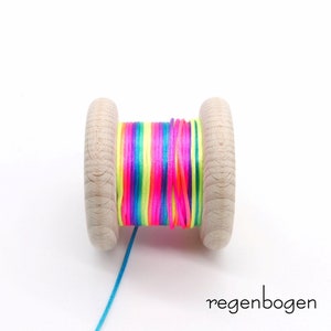 Peut inclure: Une bobine en bois enroul&eacute;e de cordon aux couleurs n&eacute;on vives, notamment rose, jaune et bleu. La bobine pr&eacute;sente une finition en bois clair naturel aux deux extr&eacute;mit&eacute;s. Le texte "regenbogen" est imprim&eacute; en noir dans le coin inf&eacute;rieur droit.