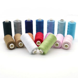 Op de afbeelding: Assortiment van 12 spoelen garen in verschillende kleuren, waaronder zwart, grijs, blauw, rood, lichtblauw, roze en groen. De spoelen zijn in een halve cirkel op een wit oppervlak gerangschikt.