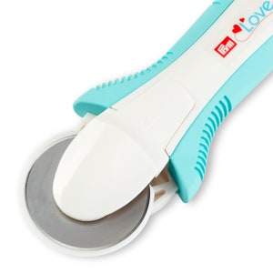 May include: A rotary cutter with a white and teal handle and a silver blade. The handle has the word "Love" and a heart design. The blade is circular and protected by a white cover.