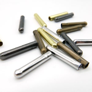 Könnte beinhalten: Eine Auswahl an Metallröhrenperlen in verschiedenen Farben, darunter Silber, Gold, Bronze und Schwarz. Die zylindrischen Perlen haben abgerundete Enden und liegen auf weißem Grund verstreut. Jede Perle ist etwa 2 cm lang.