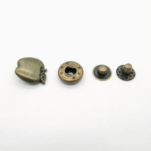 Könnte beinhalten: Vier antike, messingfarbene Druckknöpfe. Einer ist apfelförmig, der zweite rund mit Sterndetails, und die anderen beiden sind kreisförmig mit strukturierten Designs. Diese Verschlüsse werden wahrscheinlich für Kleidung oder Accessoires verwendet.