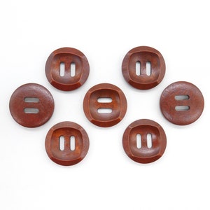 May include: Seven round, dark brown wooden buttons with two parallel rectangular holes. The buttons are arranged on a white background. The buttons have a slightly recessed centre.