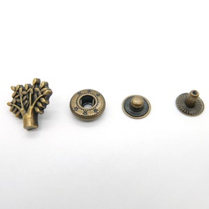 Pode incluir: Um conjunto de quatro botões de pressão de metal dourado antigo. Os botões de pressão apresentam um design de árvore, um botão redondo com um padrão de estrelas, uma base redonda e uma parte superior redonda.