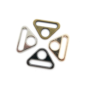 Op de afbeelding: Vier driehoekige metalen gespen in verschillende afwerkingen: zilver, goud, zwart en antiek messing. Elke gesp heeft een rechthoekige opening en een klein rond gat in het midden.