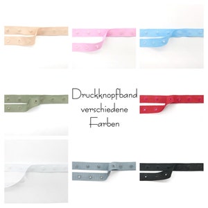 Könnte beinhalten: Eine Auswahl an farbigen Druckbandbändern in verschiedenen Farbtönen, darunter Beige, Rosa, Blau, Grün, Rot, Weiß, Grau und Schwarz. Jedes Band hat gleichmäßig verteilte Druckknöpfe. Der Text "Druckknopfband verschiedene Farben" ist ebenfalls sichtbar.