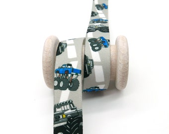 May include: Light grey ribbon with a repeating pattern of monster trucks in blue, black and grey, driving on a road. The ribbon is wrapped around a light wooden spool. The design is clearly visible.