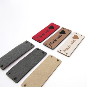 Op de afbeelding: Vier leren labels met de tekst "Made with" en een hartvormig ontwerp. De labels zijn in rood, bruin, beige en wit.
