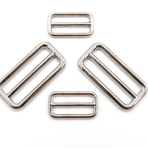 Op de afbeelding: Vier zilveren metalen schuifgespen in verschillende maten. De gespen zijn rechthoekig met twee parallelle balken in het midden.