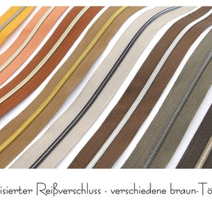 Op de afbeelding: Assortiment van ritsbanden in verschillende bruintinten met metallic accenten. De ritsbanden zijn diagonaal gerangschikt. "metallisierter Reißverschluss - verschiedene braun-Töne" staat in zwarte tekst onderaan de afbeelding.