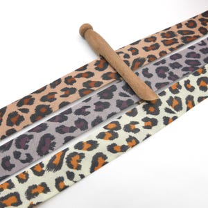 Könnte beinhalten: Drei Stoffbänder mit Leopardenmuster in den Farbtönen Beige, Grau und Creme, mit einem Holzwerkzeug darauf. Die Bänder haben schwarze und orangefarbene Flecken. Das Holzwerkzeug ist hellbraun.