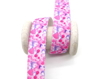 May include: A wooden spool holding a ribbon patterned with pink, purple, and red hearts. The ribbon is unwound, revealing the heart pattern. The background is plain white.