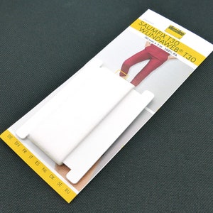 Könnte beinhalten: Eine Packung Saumfix T30 Wundaweb T30, ein weißes Bügelband, liegt auf einer dunkelgrünen, strukturierten Oberfläche. Die Verpackung enthält Text in mehreren Sprachen und eine Illustration einer Hose, die gesäumt wird.