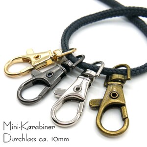 May include: Four mini-carabiners in different finishes: gold, black, silver, and bronze. The carabiners are attached to a black cord. Mini-Karabiner Durchlass ca. 10mm.