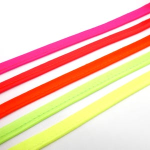Könnte beinhalten: Nahaufnahme von vier Stoffstreifen in Neonfarben. Die Streifen sind diagonal angeordnet und zeigen eine lebendige Palette aus Pink, Rot, Orange und Gelbgrün. Jeder Streifen hat einen genähten Rand, was auf ein fertiges Produkt hindeutet.