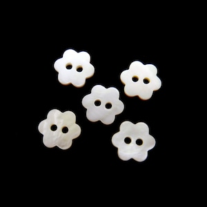 Peut inclure: Cinq boutons en forme de fleur, chacun percé de deux trous. Les boutons sont blancs avec un éclat nacré, sur fond noir. Ces boutons décoratifs sont parfaits pour la couture et les travaux manuels.