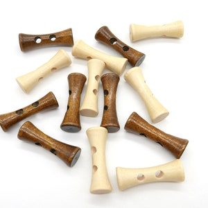 Könnte beinhalten: Eine Sammlung von Holzknebelknöpfen in verschiedenen Braun- und Naturbeigetönen. Jeder Knopf hat eine charakteristische Sanduhrform mit zwei Löchern zum Annähen. Die Knöpfe liegen auf einer weißen Oberfläche verstreut.