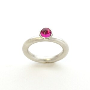 Könnte beinhalten: Ein silberner Ring mit einem polierten, magentafarbenen Edelstein. Der Ring hat ein schlichtes, minimalistisches Design mit einer glatten, matten Oberfläche. Der Edelstein ist in einer Lünettenfassung gefasst.