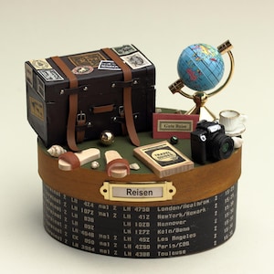Könnte beinhalten: Eine Miniatur-Holzkiste mit braunem Deckel und einem goldenen Schild mit der Aufschrift "Reisen". Die Kiste ist gefüllt mit Miniatur-Reiseartikeln, darunter ein Koffer, ein Globus, eine Kamera, ein Buch und ein Paar Sandalen. Die Kiste hat auch eine Liste von Zielen und Flugnummern an der Seite.