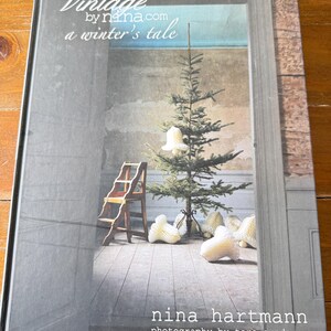 Könnte beinhalten: Ein Hardcover-Buch mit dem Titel "Vintage by nina.com a winter's tale". Das Cover zeigt einen Weihnachtsbaum mit Ornamenten, eine kleine Holzleiter und dekorative Objekte. Autorin ist Nina Hartmann, Fotos von Tore Sundgren.