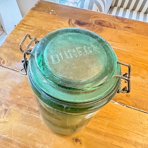 Op de afbeelding: Een groene glazen pot met een metalen deksel en een roestige metalen sluiting. De pot heeft het woord "DUMMER" op het deksel.