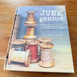 Op de afbeelding: Een boek getiteld "JUNK genius" met de ondertitel "Stylish ways to repurpose everyday objects, with over 80 projects and ideas." De cover bevat illustraties van houten spoelen en een houten object. Het boek ligt op een houten oppervlak.