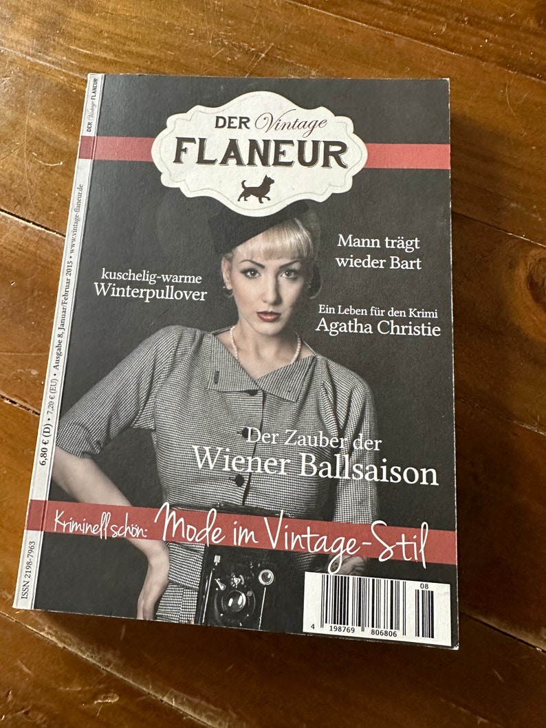 May include: A magazine titled "DER Vintage FLANEUR" featuring a woman in vintage attire. The cover includes text in German, with headlines like "kuschelig-warme Winterpullover" and "Mode im Vintage-Stil". The magazine is on a wooden surface.