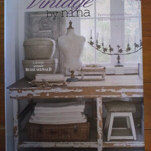 Könnte beinhalten: Buchcover "Vintage by Nina" mit Vintage-Ambiente. Es zeigt eine Schaufensterpuppe, gestapelte Behälter und einen Kerzenleuchter auf einem rustikalen Tisch. Der Untertitel des Buches lautet "Persönlich einrichten mit antiken Schätzen."