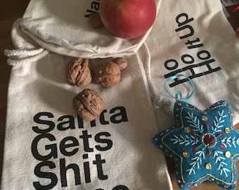 May include: Two white fabric bags with drawstring closures. One bag has the text "Naughty > Nice" printed on it and the other bag has the text "Ho Ho Ho It Up" printed on it. The bag with the text "Naughty > Nice" also has the text "Santa Gets Shit Done" printed on it. There is a red apple and some walnuts on the bag with the text "Naughty > Nice". A blue star-shaped ornament is on the bag with the text "Ho Ho Ho It Up".