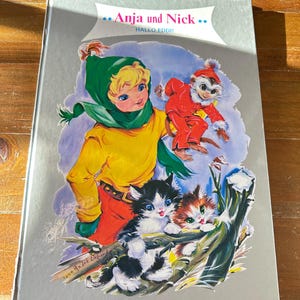 May include: A silver book cover with a colorful illustration of a girl, a monkey, and two kittens. The title of the book is "Anja und Nick" with the subtitle "Hallo Eddi!"