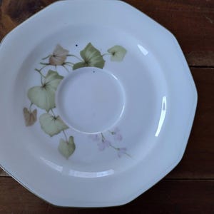 Könnte beinhalten: Weiße Untertasse mit grünem und braunem Blumendesign. Die Untertasse hat einen gewellten Rand und eine erhöhte Mitte.