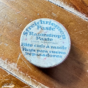 Puede incluir: Una lata redonda blanca con letras negras. La lata está etiquetada "Freichriemen Paste Razorstrope Paste Made in Germany Pâte cuir à rasoir Pasta para cueros zepasadores".