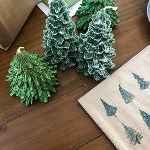 May include: Green Christmas tree-shaped candles with a textured, frosted appearance. The candles are arranged on a wooden surface, alongside a napkin with tree designs. The candles vary in size and shape, creating a festive display.