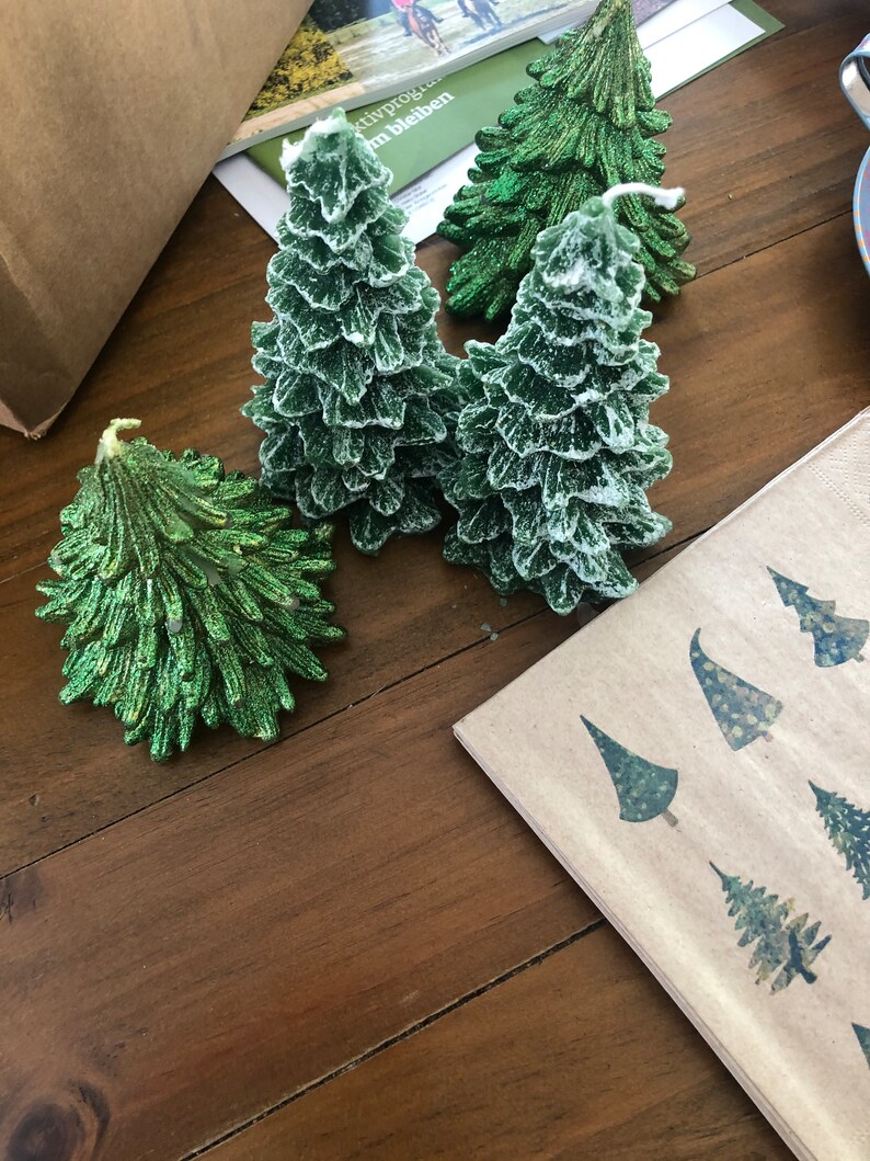 May include: Green Christmas tree-shaped candles with a textured, frosted appearance. The candles are arranged on a wooden surface, alongside a napkin with tree designs. The candles vary in size and shape, creating a festive display.