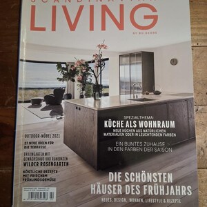 May include: A magazine cover titled "SCANDINAVIAN LIVING" with a kitchen interior design theme. The cover features a modern kitchen with a dark wood island, a vase of flowers, and text in German. Additional text includes "Outdoor Furniture 2021" and recipes.