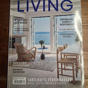 May include: A magazine cover titled "Scandinavian Living" features a bright interior with a view of the sea. The cover includes text in German, showcasing articles on architecture, design, and recipes. A wicker chair, table, and dining set are visible.