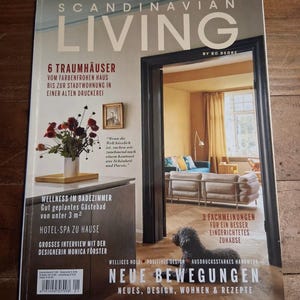 May include: A magazine cover titled "SCANDINAVIAN LIVING" with text in German. The cover features a doorway revealing a living room scene. A black dog is in the foreground. Additional text includes "6 Dream Houses" and "New Trends".
