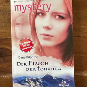 May include: A book cover with a woman's face on the front. The title is "Der Fluch Der Tohunga" and the author is Dana Kilborne. The book is part of the "mystery" series.
