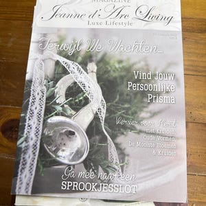 May include: A magazine cover with a white background and the title "Jeanne d'Are Living Luxe Lifestyle" in black text. The cover features a vintage-style trumpet with a white lace ribbon and greenery. The text "Terwijl We Wachten..." is in a large font at the top of the cover. The text "Vind Jouw Persoonlijke Prisma" is in a smaller font below the trumpet. The text "Versier voor Kerst met Kruiden, Oude Vormen, De Mooiste Bloemen & Kruiden" is in a smaller font below the Prisma text. The text "Ga mee naar een SPROOKJESSLOT" is in a large font at the bottom of the cover.
