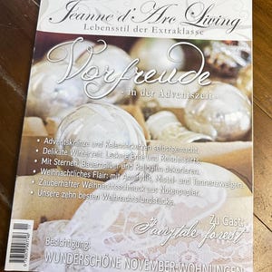 May include: A magazine cover with a white background and the title "Jeanne d'Arc Living" in black text. The subtitle is "Lebensstil der Extraklasse" in smaller black text. The cover features a large white text overlay that says "Vorfreude" with the subtitle "in der Adventszeit". The cover features a white background with a decorative design of white ornaments and a white lace tablecloth.