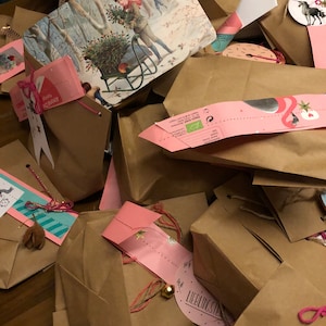 May include: A pile of brown paper gift bags, some with pink tags and string. One bag has a vintage winter scene illustration. Some tags have text and small bells. The bags are tied with pink and brown string.