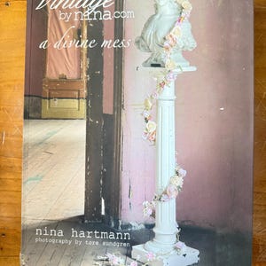 Könnte beinhalten: Ein Buch mit dem Titel "Vintage by nina.com a divine mess" mit einer weißen Büste, die mit einem Blumenkranz und einer Girlande geschmückt ist. Das Buchcover hat einen rosa-braunen Hintergrund mit dem Namen der Autorin, Nina Hartmann, und dem Fotografen, Tore Sundgren.