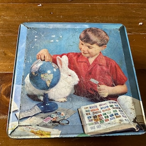 Può includere: Una vintage lunchbox di latta con un'immagine colorata di un giovane ragazzo che tiene un globo terrestre e un coniglio bianco. Il ragazzo indossa una camicia rossa e la lunchbox ha uno sfondo blu. La lunchbox presenta anche una collezione di francobolli.