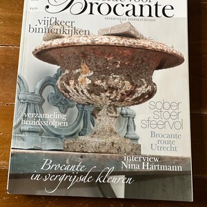 Könnte beinhalten: Das Magazin "Liefde voor Brocante" mit einem verwitterten Urnenmotiv auf dem Cover. Der Titel enthält niederländische Texte wie "vijf keer binnenkijken" und "Brocante in vergrijsde kleuren." Das Magazin hat eine rustikale, Vintage-Ästhetik.