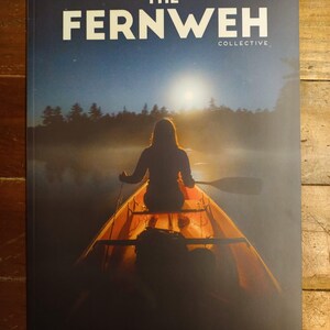 Könnte beinhalten: Magazin-Cover mit einer Person in einem Kanu auf einem nebligen See in der Abenddämmerung. Der Magazintitel lautet "THE FERNWEH COLLECTIVE" in großen weißen Buchstaben. Das Bild weckt ein Gefühl von Abenteuer und Ruhe.