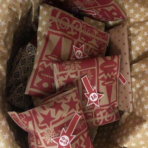 May include: A wicker basket filled with Advent calendar bags. The bags are brown with a red holiday-themed pattern and star-shaped number tags. The basket is lined with star-patterned paper.