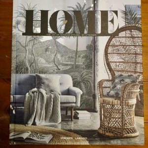 Könnte beinhalten: Ein Einband eines Wohnzeitschrift mit dem Wort "HOME" in großen schwarzen Buchstaben. Das Cover zeigt ein graues Sofa, einen gewebten Pfauenstuhl und eine Tapete mit tropischer Landschaft. Der Text "THE JOY OF INTERIOR STYLING BY VTWONEN" ist ebenfalls sichtbar.