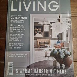 May include: A magazine cover titled "SCANDINAVIAN LIVING" with the text "5 WARME HÄUSER MIT HERZ". The cover features a room interior with a modern chandelier, a sofa, and a table with a floral arrangement. The magazine is on a wooden surface.